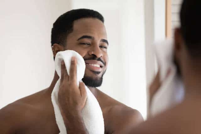 Happy handsome African American guy drying face with white towel in bathroom, looking at mirror. Young man with bare muscular shoulders and stylish beard enjoying bath routine. Grooming concept